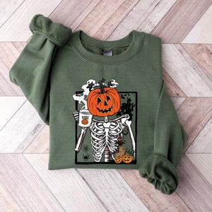 K&ouml;nnte beinhalten: Gr&uuml;nes Sweatshirt mit einem Grafikdruck eines Skeletts mit einem K&uuml;rbis-Kopf, das eine Tasse h&auml;lt, auf der "Happy Halloween" steht.