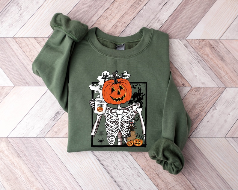 K&ouml;nnte beinhalten: Gr&uuml;nes Sweatshirt mit einem Grafikdruck eines Skeletts mit einem K&uuml;rbis-Kopf, das eine Tasse h&auml;lt, auf der "Happy Halloween" steht.
