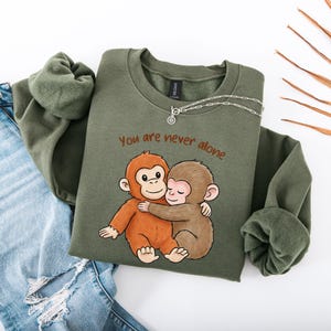 Puede incluir: Una sudadera verde oliva con el texto "You are never alone" y un gr&aacute;fico de dos monos abraz&aacute;ndose. Un collar plateado est&aacute; sobre la sudadera. La sudadera se combina con unos vaqueros azules.