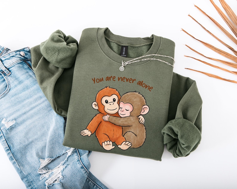 Puede incluir: Una sudadera verde oliva con el texto "You are never alone" y un gr&aacute;fico de dos monos abraz&aacute;ndose. Un collar plateado est&aacute; sobre la sudadera. La sudadera se combina con unos vaqueros azules.