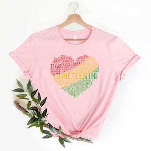 May include: A light pink t-shirt featuring a heart-shaped design composed of words related to Juneteenth. The design includes words like "Juneteenth," "Freedom," "Equality," and "Texas" in red, yellow, and green.