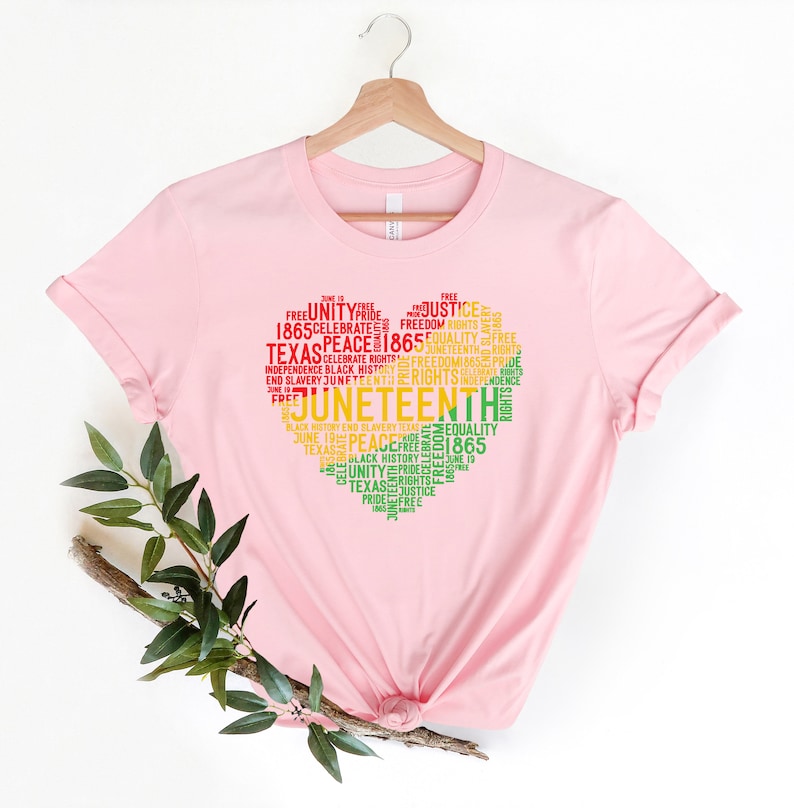 May include: A light pink t-shirt featuring a heart-shaped design composed of words related to Juneteenth. The design includes words like "Juneteenth," "Freedom," "Equality," and "Texas" in red, yellow, and green.