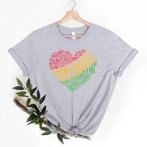 May include: A light gray t-shirt with a heart-shaped design filled with words in red, yellow, and green. The words include "Juneteenth," "Unity," "Freedom," and "1865." The shirt is hanging on a wooden hanger.