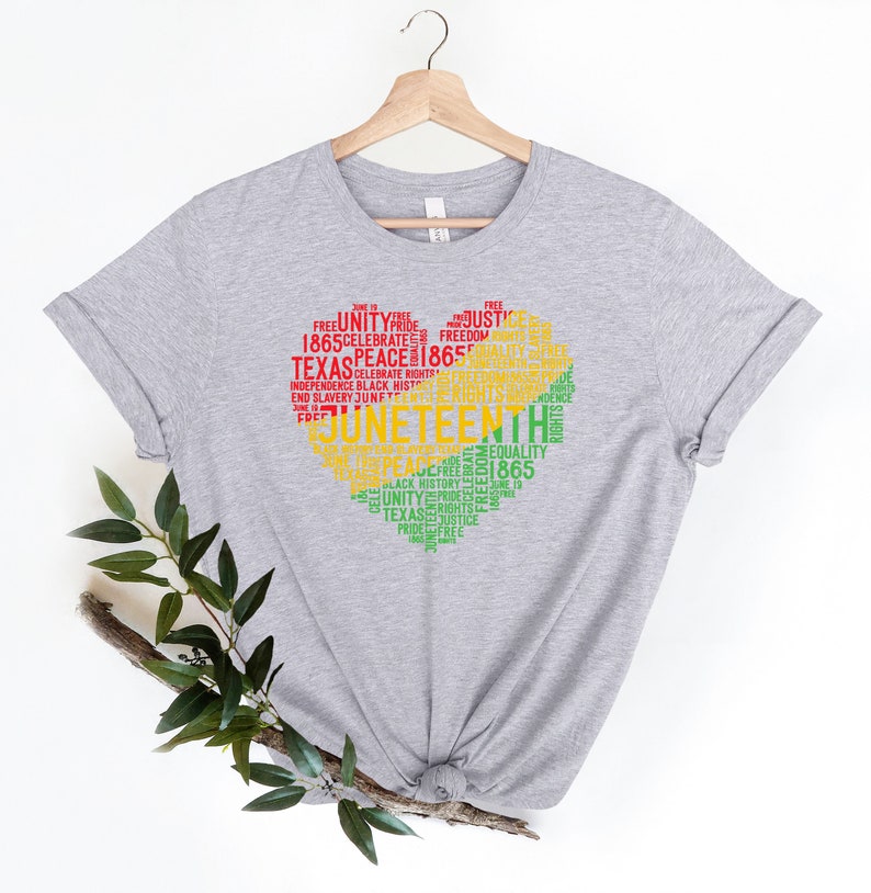 May include: A light gray t-shirt with a heart-shaped design filled with words in red, yellow, and green. The words include "Juneteenth," "Unity," "Freedom," and "1865." The shirt is hanging on a wooden hanger.
