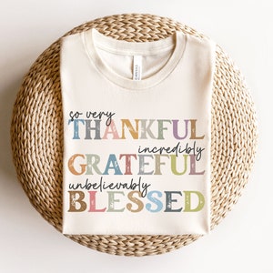 K&ouml;nnte beinhalten: Ein wei&szlig;es T-Shirt mit dem Text "so very THANKFUL incredibly GRATEFUL unbelievably BLESSED" in bunten, abgenutzten Buchstaben.