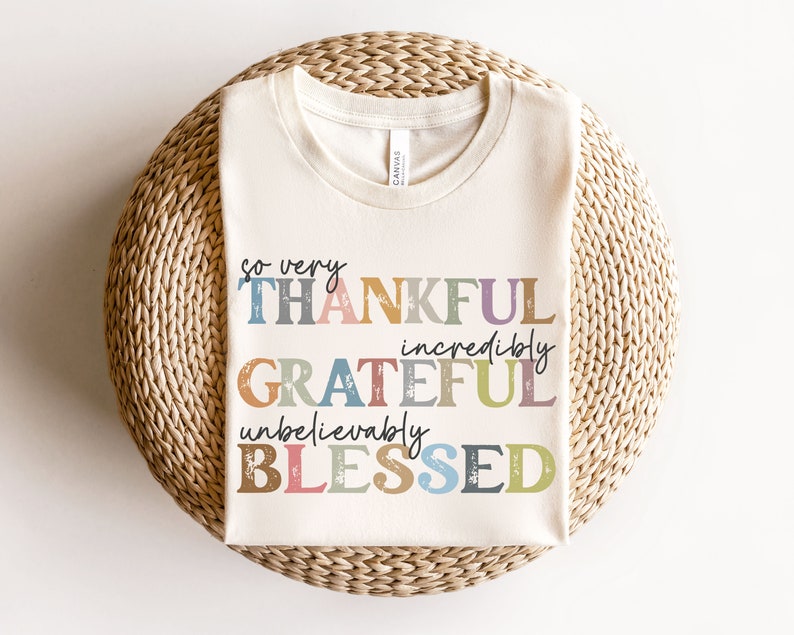 K&ouml;nnte beinhalten: Ein wei&szlig;es T-Shirt mit dem Text "so very THANKFUL incredibly GRATEFUL unbelievably BLESSED" in bunten, abgenutzten Buchstaben.