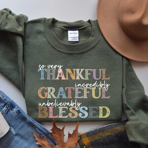 K&ouml;nnte beinhalten: Ein gr&uuml;ner Pullover mit dem Text "so very THANKFUL incredibly GRATEFUL unbelievably BLESSED" in bunten, vintage-artigen Lettern.
