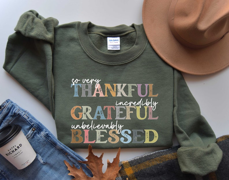 K&ouml;nnte beinhalten: Ein gr&uuml;ner Pullover mit dem Text "so very THANKFUL incredibly GRATEFUL unbelievably BLESSED" in bunten, vintage-artigen Lettern.