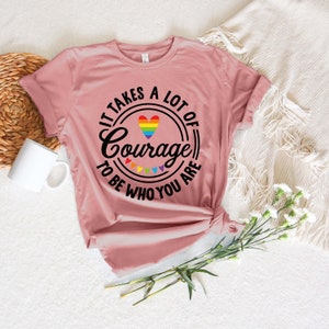May include: A dusty rose t-shirt with the text "It takes a lot of Courage to be who you are" in a black circular design. The design includes a rainbow heart and rainbow triangles. The shirt is knotted at the bottom.