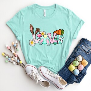 May include: A light blue t-shirt with a graphic of a bunny rabbit and the word "LOVE" in colorful letters. The letters are decorated with Easter eggs, flowers, and a carrot.