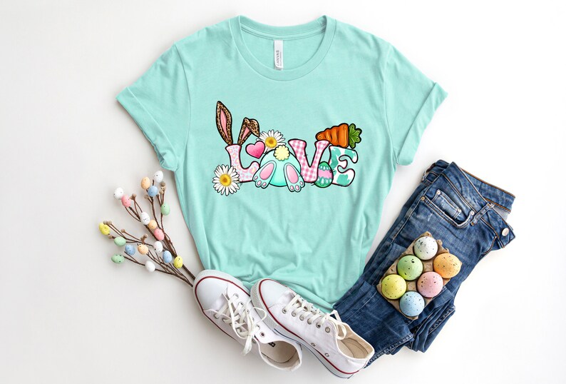 May include: A light blue t-shirt with a graphic of a bunny rabbit and the word "LOVE" in colorful letters. The letters are decorated with Easter eggs, flowers, and a carrot.
