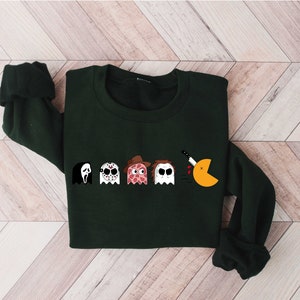 Pode incluir: Uma camisola verde escura com uma estampa gr&aacute;fica de cinco personagens fantasmas, incluindo um fantasma com uma faca, um fantasma com uma m&aacute;scara de h&oacute;quei, um fantasma com uma fatia de pizza, um fantasma com um chap&eacute;u e um fantasma com uma faca. Os fantasmas est&atilde;o em fila e o &uacute;ltimo fantasma est&aacute; comendo um Pac-Man amarelo.