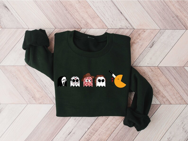 Pode incluir: Uma camisola verde escura com uma estampa gr&aacute;fica de cinco personagens fantasmas, incluindo um fantasma com uma faca, um fantasma com uma m&aacute;scara de h&oacute;quei, um fantasma com uma fatia de pizza, um fantasma com um chap&eacute;u e um fantasma com uma faca. Os fantasmas est&atilde;o em fila e o &uacute;ltimo fantasma est&aacute; comendo um Pac-Man amarelo.