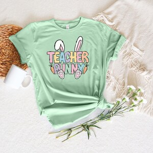 May include: A light green t-shirt with a graphic design of bunny ears, carrots, and the text "TEACHER BUNNY" in rainbow colors.