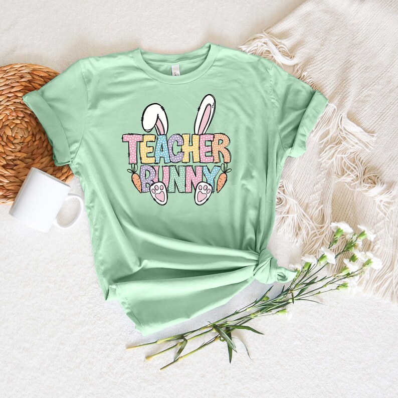 May include: A light green t-shirt with a graphic design of bunny ears, carrots, and the text "TEACHER BUNNY" in rainbow colors.