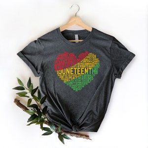 May include: Dark gray t-shirt with a heart-shaped design filled with words in red, yellow, and green. The words include "Juneteenth", "Freedom", "Equality", and "1865". The shirt is hanging on a wooden hanger.