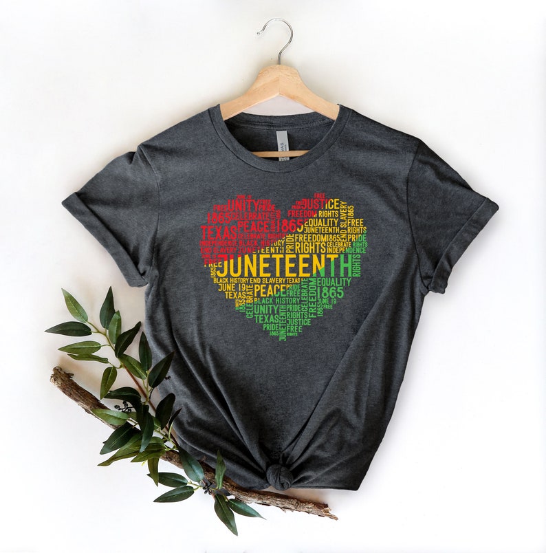 May include: Dark gray t-shirt with a heart-shaped design filled with words in red, yellow, and green. The words include "Juneteenth", "Freedom", "Equality", and "1865". The shirt is hanging on a wooden hanger.