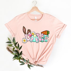 May include: A pink t-shirt with a graphic design of the word "LOVE" with Easter-themed elements, including a bunny, carrots, and eggs.