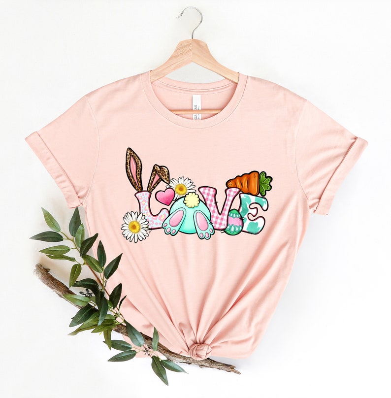 May include: A pink t-shirt with a graphic design of the word "LOVE" with Easter-themed elements, including a bunny, carrots, and eggs.