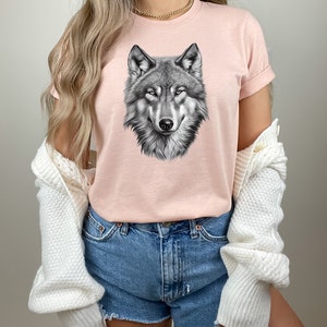 以下が含まれることがあります： 黒と白のオオカミのグラフィックプリントが施されたピンクのTシャツ。オオカミの頭は正面を向いており、口は閉じられています。Tシャツは半袖でクルーネックです。