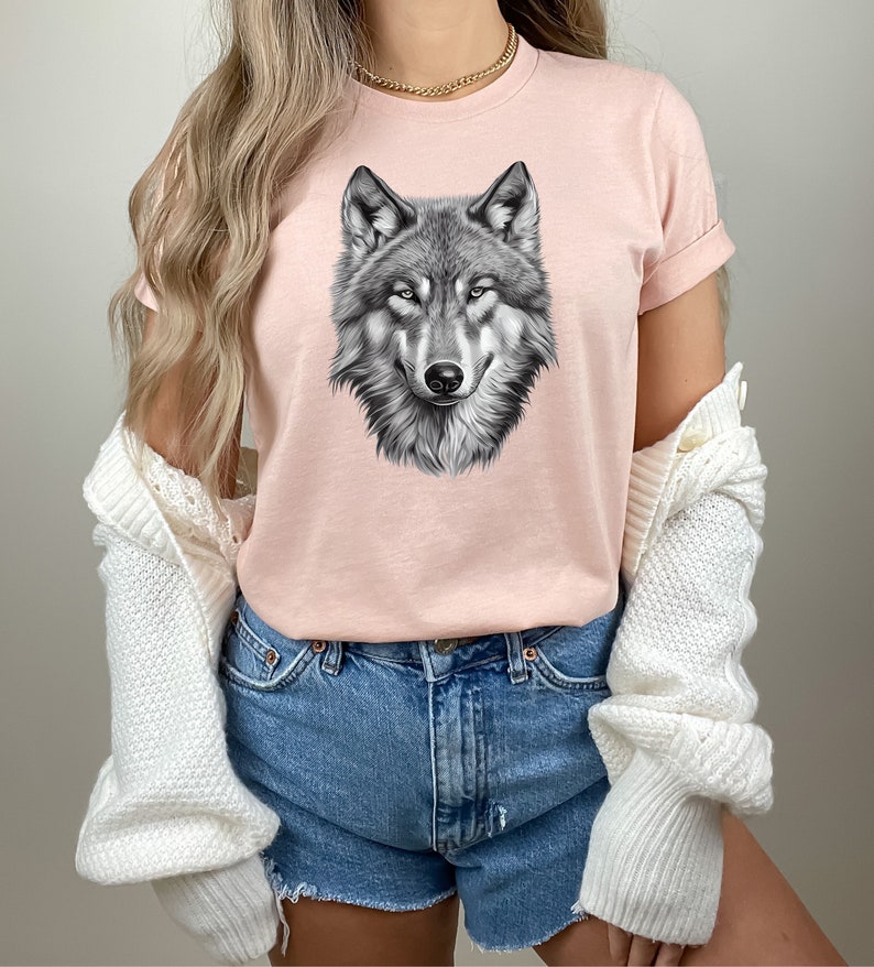以下が含まれることがあります： 黒と白のオオカミのグラフィックプリントが施されたピンクのTシャツ。オオカミの頭は正面を向いており、口は閉じられています。Tシャツは半袖でクルーネックです。