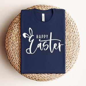 Puede incluir: Una camiseta azul marino con un dise&ntilde;o de orejas de conejo blancas y el texto "Happy Easter" en escritura blanca.