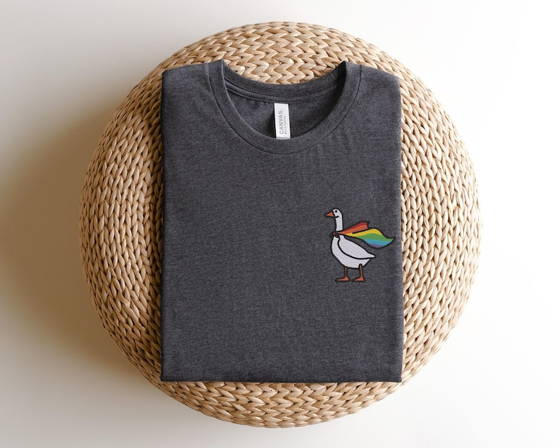 May include: A dark grey t-shirt with a white goose wearing a rainbow cape. The goose is facing right and has a small black beak. The t-shirt has a white tag that says "Canvas" on it.