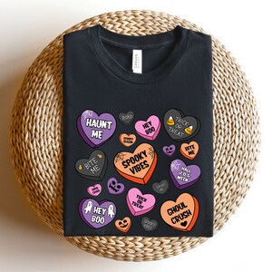 Puede incluir: Camiseta negra con un dise&ntilde;o de tem&aacute;tica de Halloween con corazones de caramelo con frases como "Haunt Me", "Spooky Vibes", "Ghoul Crush" y "Trick or Treat".