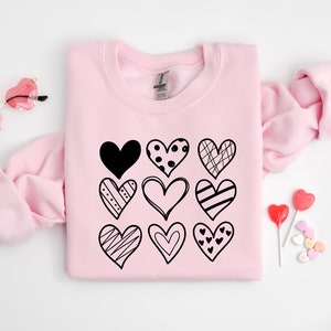 May include: Pink sweatshirt featuring a black graphic of nine different hearts. The hearts are all in different styles and have various patterns.