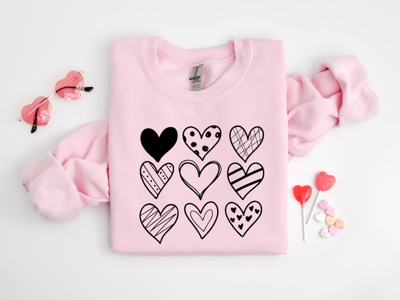 May include: Pink sweatshirt featuring a black graphic of nine different hearts. The hearts are all in different styles and have various patterns.