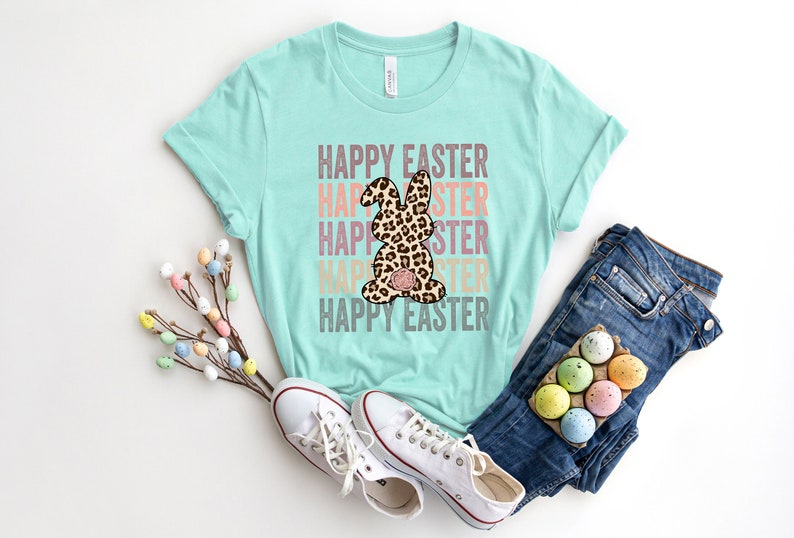 May include: A turquoise t-shirt with the text "Happy Easter" repeated in a rainbow color gradient. A leopard print bunny is centered on the shirt. The shirt is laid out on a white surface with a pair of white sneakers and blue jeans.