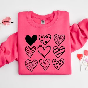 May include: A pink sweatshirt featuring a black design of nine different hearts. The hearts are all different shapes and sizes and have various patterns.