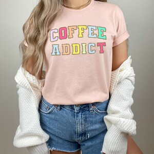 May include: A pink t-shirt with the text "COFFEE ADDICT" in colorful letters with a gold glitter outline.