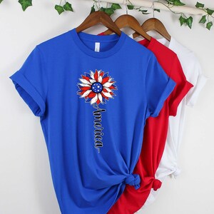 Puede incluir: Una camiseta azul, roja y blanca con un dise&ntilde;o de girasol con la bandera estadounidense. El texto "America" est&aacute; impreso verticalmente debajo del girasol.
