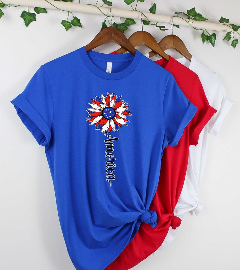 Puede incluir: Una camiseta azul, roja y blanca con un dise&ntilde;o de girasol con la bandera estadounidense. El texto "America" est&aacute; impreso verticalmente debajo del girasol.