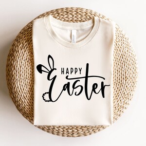 Puede incluir: Una camiseta blanca con un dise&ntilde;o de orejas de conejo negras y el texto "Happy Easter" en una fuente cursiva.