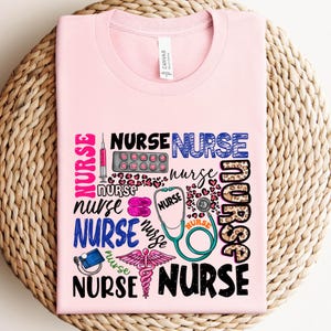May include: A light pink t-shirt with the word "NURSE" in various fonts and colors. The design includes medical symbols like a stethoscope, syringe, pills, and caduceus. The shirt is made of a soft material.