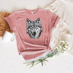 以下が含まれることがあります： ピンクのTシャツに、白黒のオオカミのグラフィックプリントが施されています。オオカミの頭は正面を向いており、口は閉じられています。