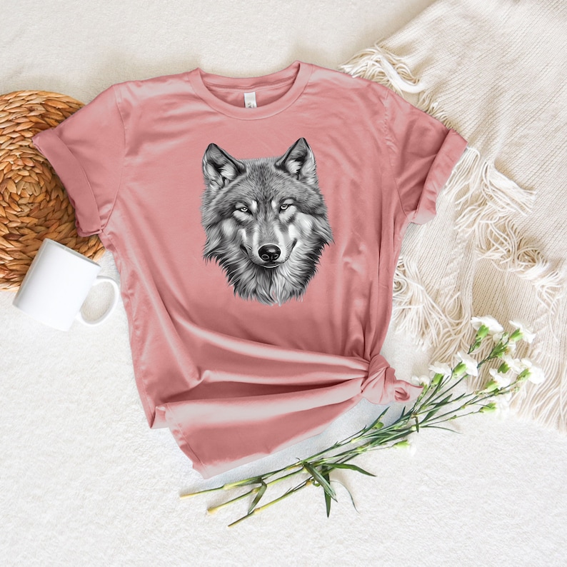 以下が含まれることがあります： ピンクのTシャツに、白黒のオオカミのグラフィックプリントが施されています。オオカミの頭は正面を向いており、口は閉じられています。