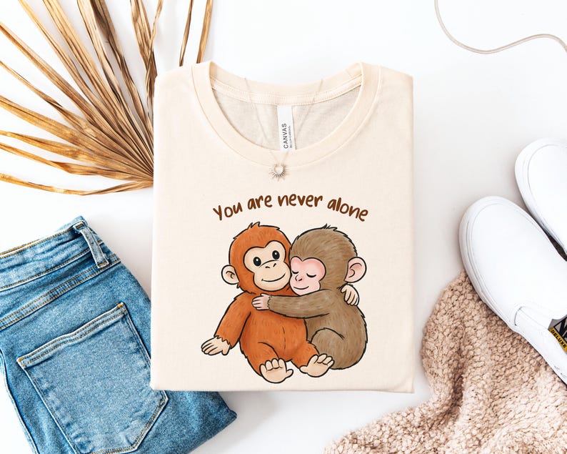 Puede incluir: Camiseta color crema con una ilustraci&oacute;n de dibujos animados de dos monos abraz&aacute;ndose. El texto "You are never alone" est&aacute; impreso encima de los monos. La camiseta est&aacute; doblada y colocada sobre una superficie blanca con vaqueros azules y zapatos blancos.