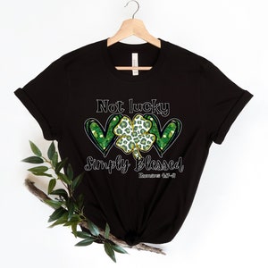 Puede incluir: Camiseta negra con un tr&eacute;bol verde y dos corazones con detalles de purpurina dorada. El texto "Not lucky simply blessed Romans 4:17-8" est&aacute; impreso en la camiseta.
