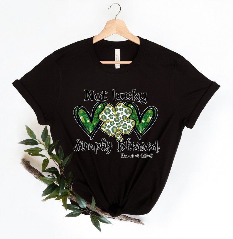 Puede incluir: Camiseta negra con un tr&eacute;bol verde y dos corazones con detalles de purpurina dorada. El texto "Not lucky simply blessed Romans 4:17-8" est&aacute; impreso en la camiseta.