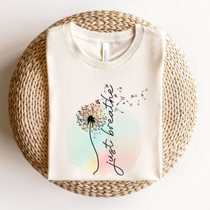 May include: Cream-coloured t-shirt with a dandelion design and the words "just breathe" in cursive. The dandelion is central, set against a pastel watercolour background. The shirt is folded on a woven surface.