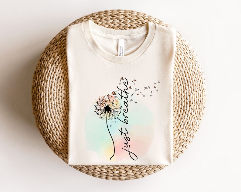 May include: Cream-coloured t-shirt with a dandelion design and the words "just breathe" in cursive. The dandelion is central, set against a pastel watercolour background. The shirt is folded on a woven surface.