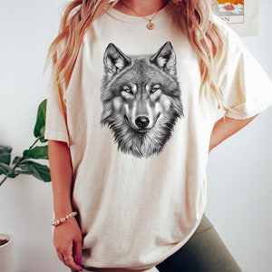 以下が含まれることがあります： 白のオーバーサイズTシャツに、白黒のオオカミのグラフィックプリントが施されています。オオカミは直接見ています。
