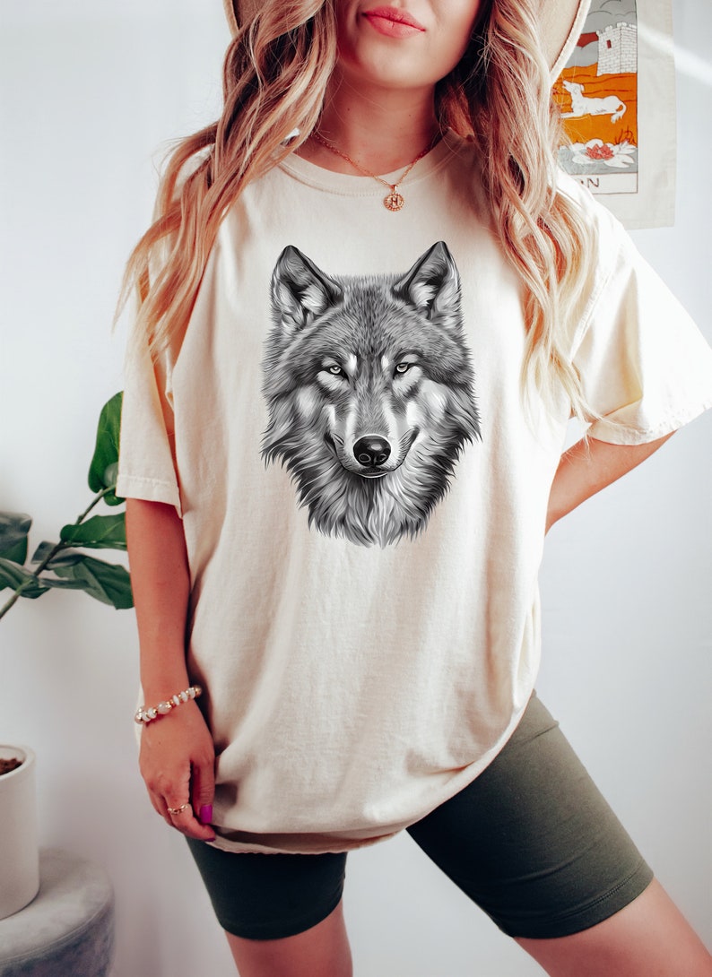 以下が含まれることがあります： 白のオーバーサイズTシャツに、白黒のオオカミのグラフィックプリントが施されています。オオカミは直接見ています。