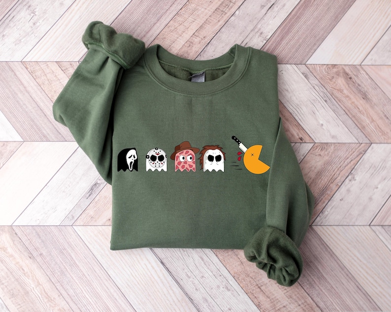 Pode incluir: Camisola verde com um gr&aacute;fico de personagens de filmes de terror como fantasmas Pac-Man. Os personagens incluem Ghostface, Jason Voorhees, Freddy Krueger e Michael Myers. O fantasma Pac-Man est&aacute; a comer uma faca.