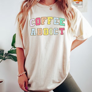 May include: A cream colored t-shirt with the text "COFFEE ADDICT" in rainbow colored letters with gold glitter outlining each letter.