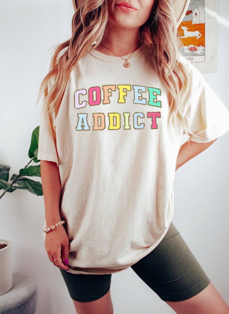 May include: A cream colored t-shirt with the text "COFFEE ADDICT" in rainbow colored letters with gold glitter outlining each letter.