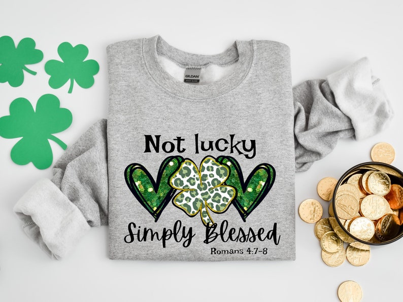 Puede incluir: Sudadera gris con un tr&eacute;bol verde y dos corazones con un dise&ntilde;o de purpurina dorada. El texto dice "Not lucky Simply Blessed Romans 4:7-8".